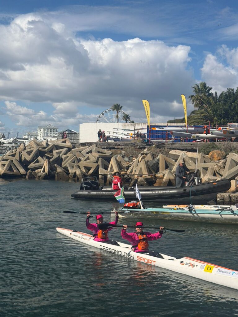 Bridget Boulle and Angela Taylor paddling into the finish of the 2026 Prescient Freedom Paddle with arms raised, the Ferris wheel and V&A Waterfront visible in the background